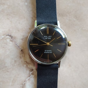 Könnte beinhalten: Eine Armbanduhr mit schwarzem Zifferblatt und goldenen Akzenten. Das Zifferblatt der Uhr trägt die Aufschrift "Poljot de Luxe" und "23 jewels shockproof". Die Uhr ist an einem schwarzen Lederarmband.