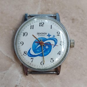 Op de afbeelding: Vintage Sekonda polshorloge met een witte wijzerplaat, zwarte cijfers en een blauw planeetontwerp. De kast en wijzers zijn zilverkleurig. De tekst "SEKONDA 19 JEWELS" is zichtbaar. Een klassiek horloge met een diameter van ongeveer 3,5 cm.