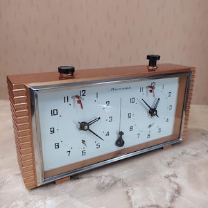 May include: A vintage chess clock with a copper-colored metal frame and two white clock faces. The clock features black hands, black knobs, and the Russian word "Янтарь" on the face. The clock is designed for competitive chess play.