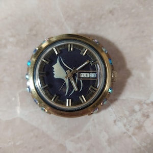 Könnte beinhalten: Eine Vintage-Armbanduhr in Goldoptik mit einem dunkelblauen Zifferblatt, das eine weiße Silhouette eines Frauenprofils zeigt. Die Uhr hat goldene Zeiger und Markierungen, eine Datumsanzeige mit "DI 23" und ist mit schillernden Kristallen um die Lünette verziert.