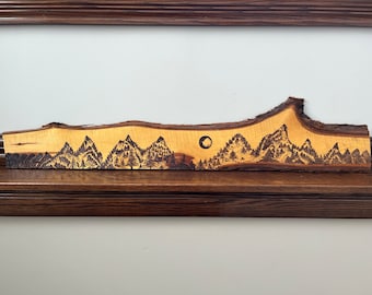 May include: A wood slice with a woodburned design of a mountain range and a full moon. The wood is a light brown colour and the design is black.