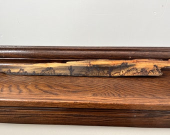 Può includere: Un primo piano di una tavola di legno con un disegno bruciato che rappresenta un paesaggio desertico. Il disegno include montagne, cactus e un tramonto.