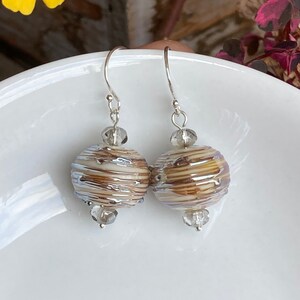 May include: A pair of handcrafted earrings featuring round, striped beads in shades of brown, cream, and silver. The earrings have silver hooks and are accented with clear, faceted beads. The earrings are displayed on a white surface.
