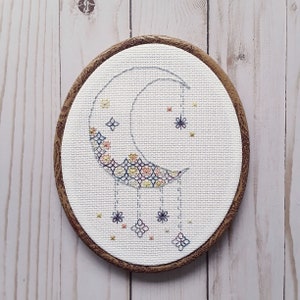 Puede incluir: Un aro de bordado blanco con un marco de madera marrón. El bordado representa una luna creciente con un patrón floral y estrellas colgantes.