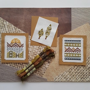May include: Three cross-stitch patterns on brown cardstock with embroidery floss. The patterns are a geometric design with a sun, a pair of trees, and a geometric pattern with mountains.