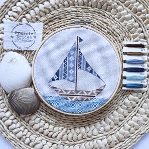 Può includere: Un disegno di barca a vela a punto croce in un telaio di legno. La barca a vela presenta motivi blu e bianchi sulla vela e sullo scafo, con un bordo d'acqua blu. L'immagine include un'etichetta Prairie Bridge Stitchery, conchiglie e filo da ricamo.