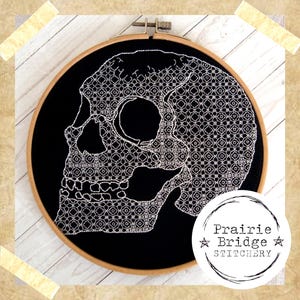 May include: White embroidered skull design on a black fabric background in a wooden embroidery hoop. The design features a detailed geometric pattern. The text "Prairie Bridge Stitchery" is visible in the lower right corner.
