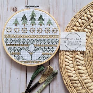May include: A completed cross-stitch embroidery in a wooden hoop. The design features green trees, snowflakes, and geometric patterns in shades of green and brown. The text "Prairie Bridge Stitchery" is visible on a small tag.
