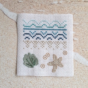 May include: A white needlepoint canvas with a beach-themed design. The top features blue and teal geometric patterns. Below are tan and brown cross-stitched elements, including a seashell, starfish, and circular designs. The canvas is on a sandy surface.