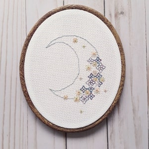 Op de afbeelding: Een houten borduurring met een witte stof eroverheen gespannen. De stof is geborduurd met een halve maanvorm en een ontwerp van kleurrijke bloemen.