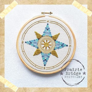 May include: Embroidered compass rose design in a wooden hoop. The compass features gold, light blue, and dark blue thread. The text "Prairie Bridge Stitchery" is in a circle at the bottom right.