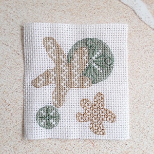 May include: A white canvas square featuring embroidered sea life designs. The design includes a starfish, a sand dollar, and a small circular shape, all in shades of tan and green. The embroidery is detailed and textured.