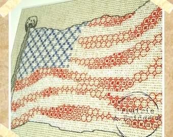 May include: A cross-stitch pattern of an American flag with red, white, and blue thread. The flag is waving in the wind. The pattern is framed with a beige border and includes the text "Prairie Bridge Stationery".