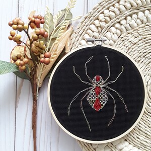 Puede incluir: Un aro de bordado negro con un diseño de araña roja y negra. La araña está bordada con hilo rojo, negro y blanco.
