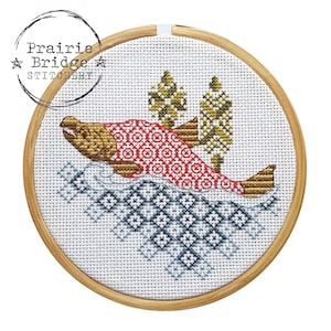 May include: A cross-stitch embroidery of a salmon jumping out of the water. The salmon is red and gold with a patterned design. The water is blue and white with a patterned design. The embroidery is framed in a round wooden hoop.