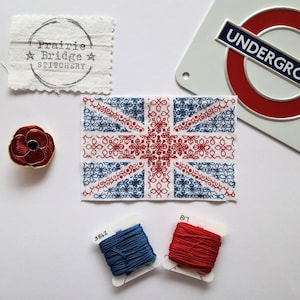 May include: A cross-stitched Union Jack flag with intricate red and blue floral designs. The image also shows a red poppy pin, spools of red and blue thread, and a "Prairie Bridge Stitchery" tag. A London Underground sign is also visible.