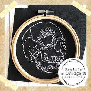 Könnte beinhalten: Ein weiß gesticktes Totenkopf-Design auf schwarzem Stoff in einem hölzernen Stickrahmen. Der Totenkopf ist mit floralen Details verziert. Der Rahmen ist mit "Prairie Bridge Stitchery" beschriftet.