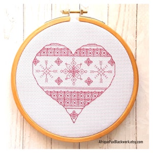 Può includere: Un ricamo a punto croce a forma di cuore rosa e bianco in un telaio da ricamo in legno. Il cuore è decorato con un motivo geometrico.