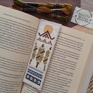 Peut inclure: Un marque-page en point de croix représentant une scène de montagne. Le marque-page présente un soleil, des montagnes, des arbres et une bordure à motifs bleus et gris. Le marque-page est réalisé avec du fil à broder coloré sur un tissu blanc.