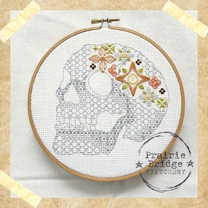 May include: Embroidered skull with floral details in shades of orange, yellow, and green. The skull is outlined in gray and the embroidery is on white fabric. The fabric is stretched over a wooden embroidery hoop. The text "Prairie Bridge Stitchery" is printed in black on the bottom right corner of the hoop.
