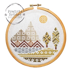 Op de afbeelding: Een kruissteekpatroon van een berglandschap met een zon, bomen en een geometrisch ontwerp. Het patroon is gestikt op witte stof en ingelijst in een houten hoepel. De tekst "Prairie Bridge Stitchery" is zichtbaar in de linkerbovenhoek.