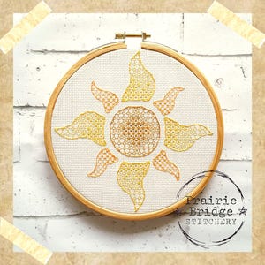 Puede incluir: Un diseño de sol bordado en amarillo y naranja en un aro de bordado de madera. El aro está en una pared de ladrillo blanco. El texto "Prairie Bridge Stitchery" está en la esquina inferior derecha.
