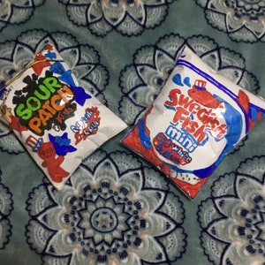 Puede incluir: Dos bolsas blancas en forma de almohada con diseños azules y rojos. La bolsa de la izquierda tiene las palabras "Sour Patch Kids" en letras azules y rojas. La bolsa de la derecha tiene las palabras "Swedish Fish Mini White Fish" en letras azules y rojas.