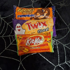 May include: Three Halloween-themed candy bar cushions. The top cushion is orange and brown with the text "Reese's Pieces Pumpkin". The middle cushion is purple and orange with the text "Twix Cookie Ghost". The bottom cushion is orange and red with the text "Kit Kat Pumpkin Pie".