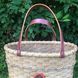 Puede incluir: Una cesta tejida con un asa de cuero marrón y un diseño circular rojo y naranja en la parte delantera.