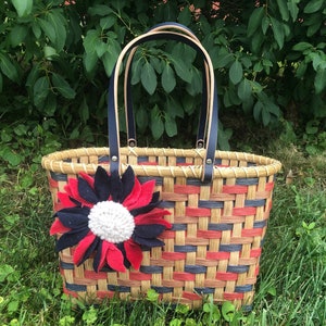 Puede incluir: Una cesta tejida con asas de cuero negro y una flor de fieltro roja, blanca y azul. La cesta está hecha de materiales naturales y tiene un patrón tejido.