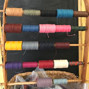May include: A wooden rack with spools of thread in various colors, including yellow, red, blue, green, and brown. The spools are arranged in rows on the rack.