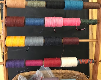 May include: A wooden rack with spools of thread in various colors, including yellow, red, blue, green, and brown. The spools are arranged in rows on the rack.