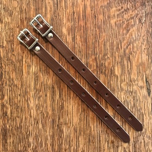 Puede incluir: Dos correas de cuero marrón con hebillas plateadas. Las correas miden aproximadamente 45 cm de largo y tienen varios agujeros para ajustar la longitud.