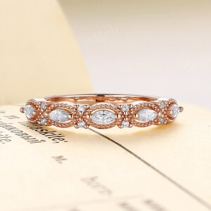 May include: Rose gold ring with a pattern of oval and round white gemstones. The ring is displayed on a vintage book page, highlighting the band's detail and the stones' sparkle. A delicate and elegant piece of jewellery.