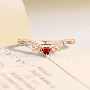 May include: Rose gold ring with a heart-shaped red gemstone at the center. The ring features a curved band with small, clear gemstones and decorative accents on either side of the heart-shaped stone. The ring is set against a vintage book page.