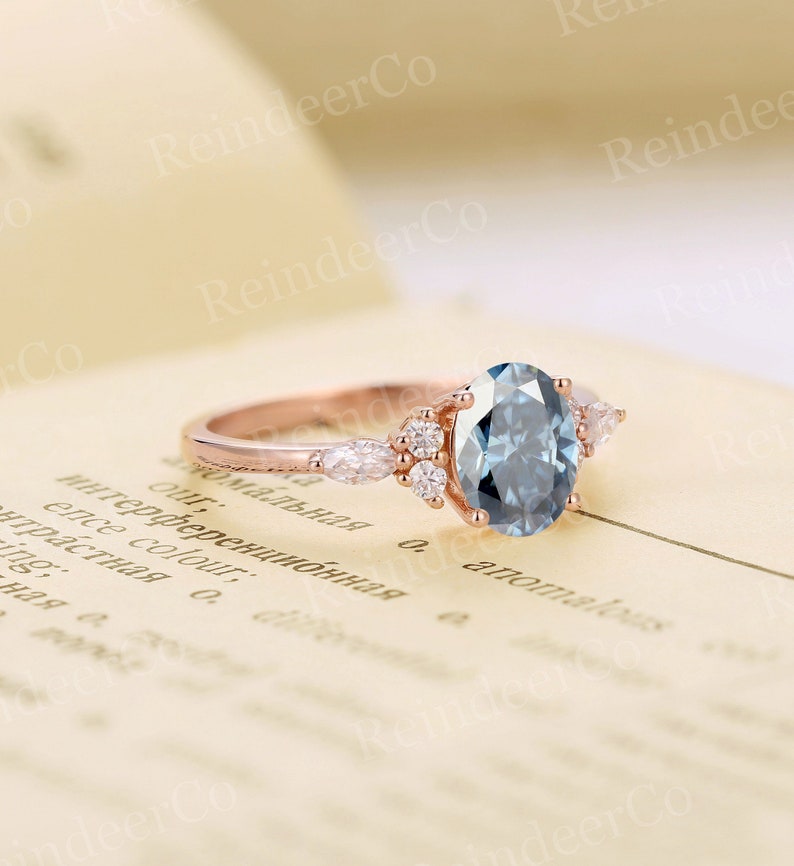 May include: A rose gold engagement ring with a large oval-shaped blue gemstone surrounded by smaller diamonds. The ring is set on a white background.