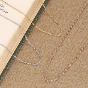 Könnte beinhalten: Drei feine Halsketten aus Gold, Silber und Roségold. Die Ketten werden auf einer strukturierten, neutral getönten Oberfläche präsentiert, mit einem Blick auf den Rand eines Buches im Hintergrund. Die Halsketten sind schlicht und elegant, für den Alltag geeignet.
