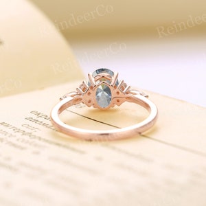 May include: A rose gold engagement ring with a light blue oval-shaped center stone and smaller accent stones. The ring is on a cream-colored surface.