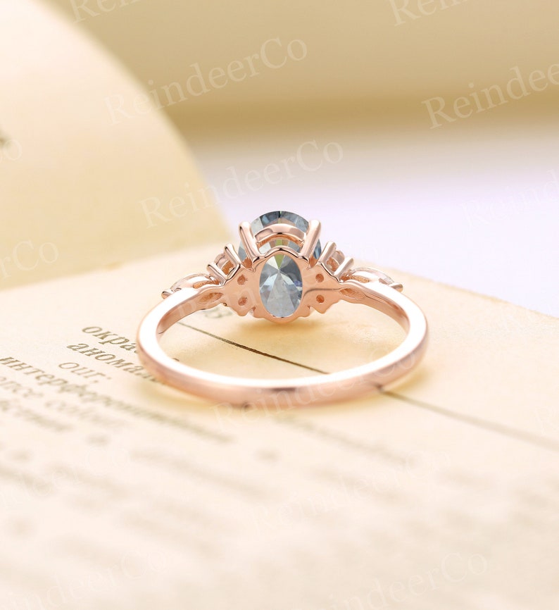 May include: A rose gold engagement ring with a light blue oval-shaped center stone and smaller accent stones. The ring is on a cream-colored surface.