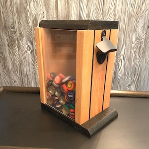 May include: A wooden bottle opener with a clear plastic container to hold bottle caps. The bottle opener is made of dark wood with a light wood trim. The container is filled with a variety of colorful bottle caps.