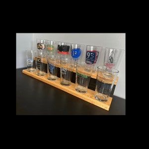 May include: A wooden beer flight with twelve clear pint glasses, each with a different logo. The glasses are arranged in two rows on a light brown wooden holder. The logos are in various colors and designs.