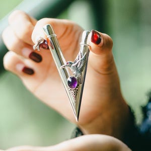 May include: A silver tie bar with a triangular design, featuring a purple gemstone and an eagle emblem. The tie bar is held in a hand with dark red nail polish, against a blurred green background.