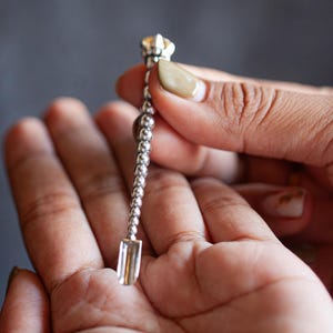May include: A silver-toned metal tool featuring a long, slender handle composed of a series of small, spherical beads. The top of the handle is capped with a decorative, faceted clear crystal, while the bottom end terminates in a flat, rectangular scoop.