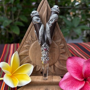 May include: A silver and wood pendant with a clear crystal point, set against a wooden sunburst design. The pendant features ornate silver detailing and a small red gemstone. A yellow and white flower and a pink flower are in the foreground.