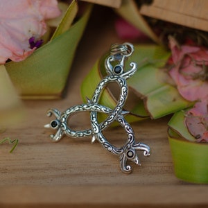 May include: A silver pendant with a three-part design featuring intricate details and black gemstones. The pendant is on a wooden surface with green leaves.
