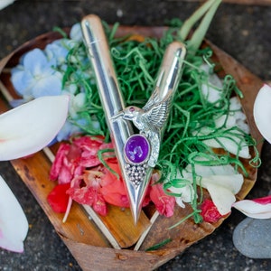 Puede incluir: Un abrecartas plateado con un dise&ntilde;o de p&aacute;jaro y una piedra preciosa p&uacute;rpura. El abrecartas descansa sobre un lecho de vegetaci&oacute;n, flores y una bandeja de madera. Flores blancas y amarillas en primer plano.