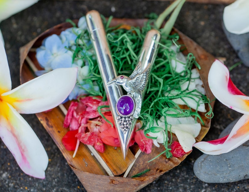 Puede incluir: Un abrecartas plateado con un dise&ntilde;o de p&aacute;jaro y una piedra preciosa p&uacute;rpura. El abrecartas descansa sobre un lecho de vegetaci&oacute;n, flores y una bandeja de madera. Flores blancas y amarillas en primer plano.