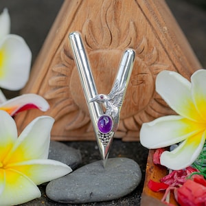 Puede incluir: Un bol&iacute;grafo plateado con un dise&ntilde;o de p&aacute;jaro y piedra preciosa p&uacute;rpura. El bol&iacute;grafo se encuentra sobre un fondo de flores blancas y amarillas, una talla de madera y piedras grises. El dise&ntilde;o del bol&iacute;grafo incluye un p&aacute;jaro con un ojo de piedra roja y una gran piedra preciosa p&uacute;rpura.