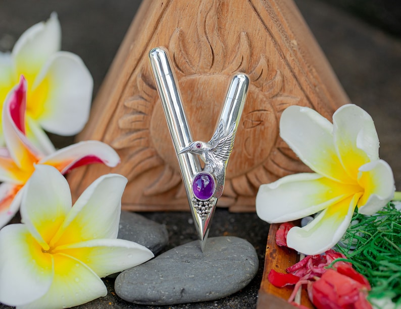 Puede incluir: Un bol&iacute;grafo plateado con un dise&ntilde;o de p&aacute;jaro y piedra preciosa p&uacute;rpura. El bol&iacute;grafo se encuentra sobre un fondo de flores blancas y amarillas, una talla de madera y piedras grises. El dise&ntilde;o del bol&iacute;grafo incluye un p&aacute;jaro con un ojo de piedra roja y una gran piedra preciosa p&uacute;rpura.