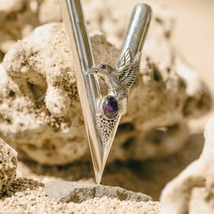 Puede incluir: Una horquilla de metal plateado con un detalle de p&aacute;jaro y piedra preciosa p&uacute;rpura. La horquilla tiene forma de V con un extremo puntiagudo y una parte superior redondeada. El p&aacute;jaro tiene las alas extendidas y est&aacute; posado sobre la piedra preciosa.